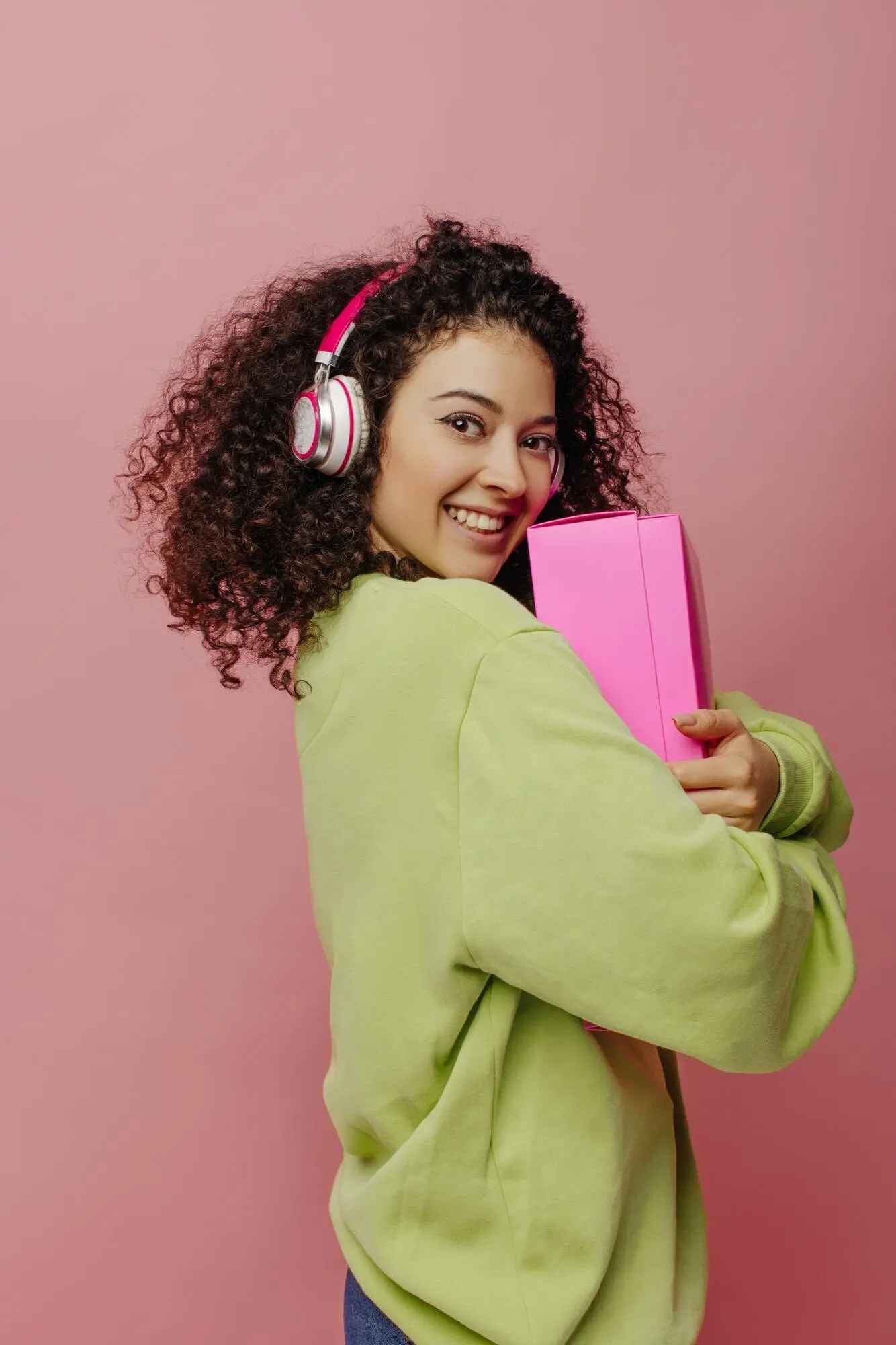 Attraktive, hellhäutige junge Frau steht seitlich und hält eine Schachtel an sich, vor pinkem Hintergrund. Die Brünette mit Kopfhörern und Jacke lächelt in die Kamera. Emotionskonzept, Gesichtsausdruck.